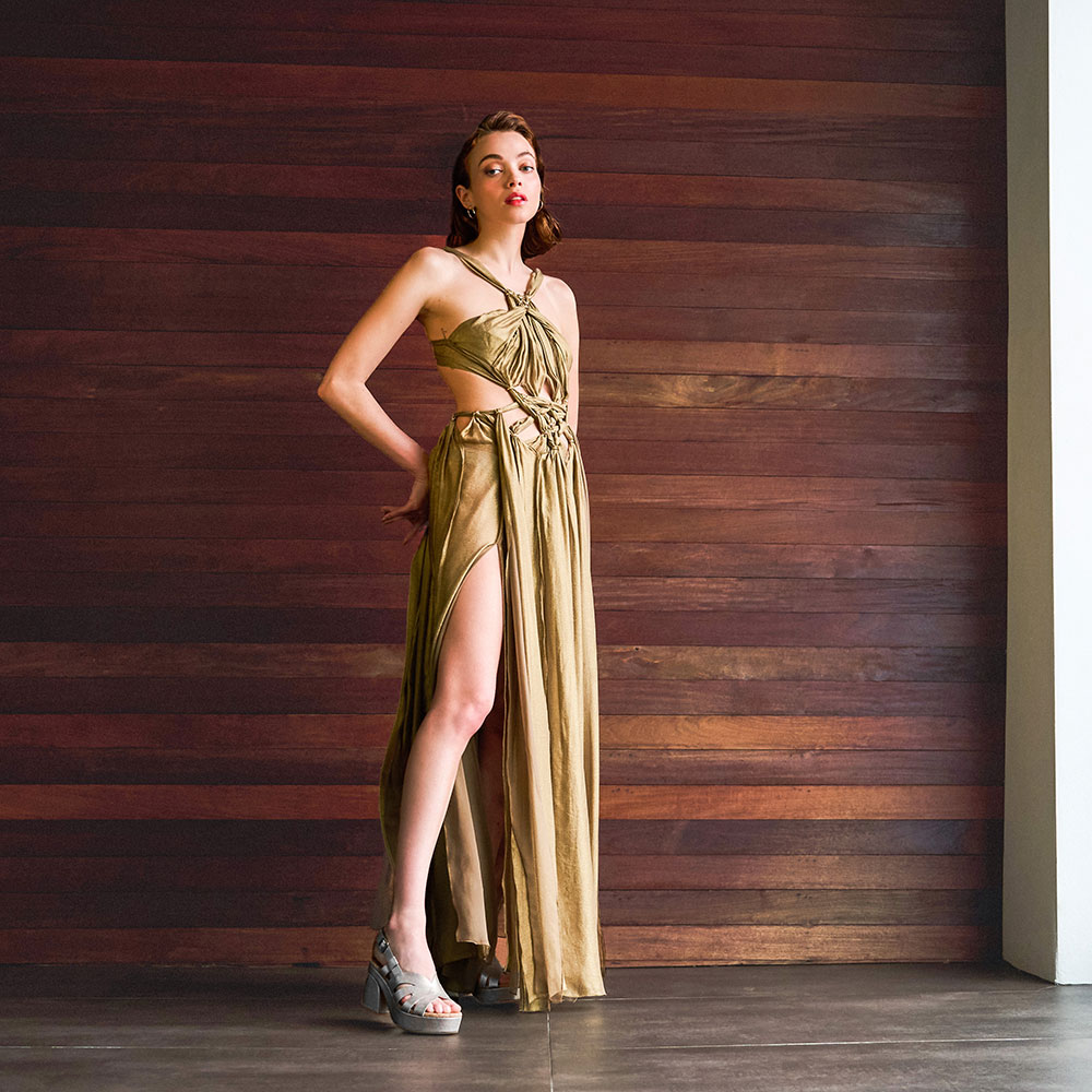 A woman standing in front of a window wearing the Paschal heeled sandal in gold metallic.